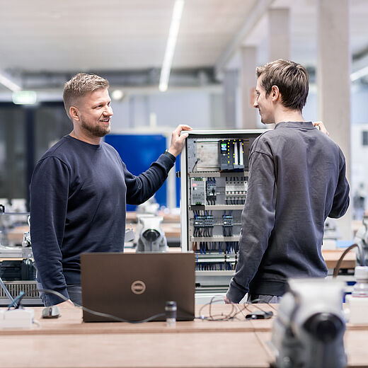 Zwei Männer stehen in einer modernen Werkstatt und betrachten gemeinsam ein Steuerungssystem. Einer zeigt auf das Gerät, während der andere aufmerksam zuhört. Auf einem Tisch im Vordergrund steht ein Laptop.