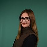 Porträt einer jungen Frau mit langen, glatten Haaren und einer Brille. Sie trägt einen braunen Pullover mit hohem Kragen und steht seitlich zur Kamera, mit verschränkten Armen. Der Hintergrund ist ein einfarbiges Grün.