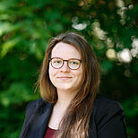 Eine lächelnde Frau mit langen, braunen Haaren und einer Brille, die in einem schwarzen Blazer gekleidet ist, steht vor einer grünen, verschwommenen Hintergrundkulisse. Sie wirkt freundlich und selbstbewusst.