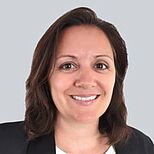 Porträt einer Frau mit dunklen, schulterlangen Haaren. Sie trägt ein schwarzes Jackett und ein helles Oberteil. Die Person lächelt freundlich in die Kamera, vor einem grauen Hintergrund.