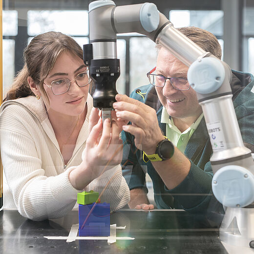 Eine junge Frau und ein älterer Mann arbeiten gemeinsam an einem Roboterarm. Beide beobachten aufmerksam, wie der Roboter einen farbigen Block greift. Die Atmosphäre ist kollegial und lehrreich, wodurch die Interaktion zwischen Mensch und Maschine betont wird.