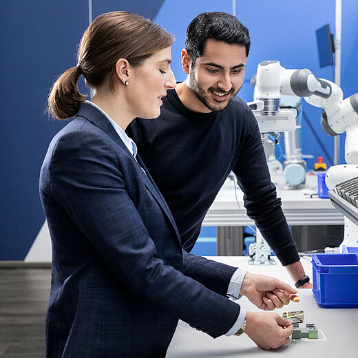 Zwei Personen stehen an einem Tisch in einem modernen Arbeitsumfeld. Eine Frau hält eine Leiterplatte in der Hand, während ein Mann interessiert zusieht. Im Hintergrund sind technische Geräte und ein Roboterarm zu erkennen.