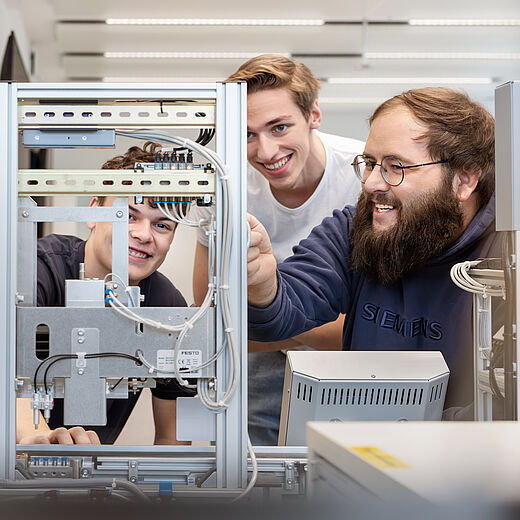 Drei junge Männer stehen an einem technischen Geräteaufbau und betrachten ihn aufmerksam. Einer von ihnen lächelt und zeigt auf eine Komponente. Die Atmosphäre wirkt kollegial und lehrreich, während sie gemeinsam an dem Projekt arbeiten.