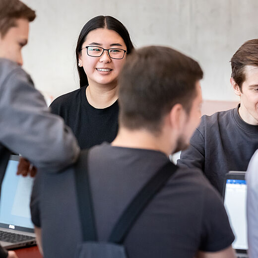Eine Gruppe junger Menschen sitzt an einem Tisch und diskutiert. Im Vordergrund ist ein Mädchen mit Brille zu sehen, das freundlich lächelt. Die anderen Teilnehmer sind teilweise abgewandt und scheinen interessiert zu sein. Auf dem Tisch befinden sich Laptops.