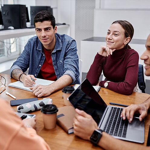 Eine Gruppe von vier Personen sitzt an einem Tisch und diskutiert. Zwei Personen haben Laptops vor sich, während eine andere Notizen macht. Alle scheinen engagiert und freundlich miteinander zu interagieren. Der Raum wirkt modern und fördert die Zusammenarbeit.