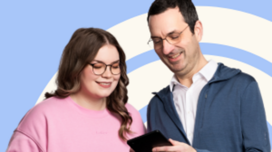 Zwei Personen stehen nebeneinander und schauen auf digitale Geräte. Eine Frau mit Brille trägt einen rosa Pullover und hält ein Tablet. Ein Mann mit Brille trägt einen blauen Hoodie und ein Hemd. Beide lächeln und scheinen sich über den Inhalt der Geräte auszutauschen.