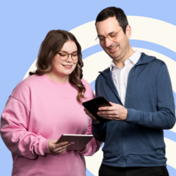 Zwei Personen stehen nebeneinander und schauen auf digitale Geräte. Eine Frau mit Brille trägt einen rosa Pullover und hält ein Tablet. Ein Mann mit Brille trägt einen blauen Hoodie und ein Hemd. Beide lächeln und scheinen sich über den Inhalt der Geräte auszutauschen.