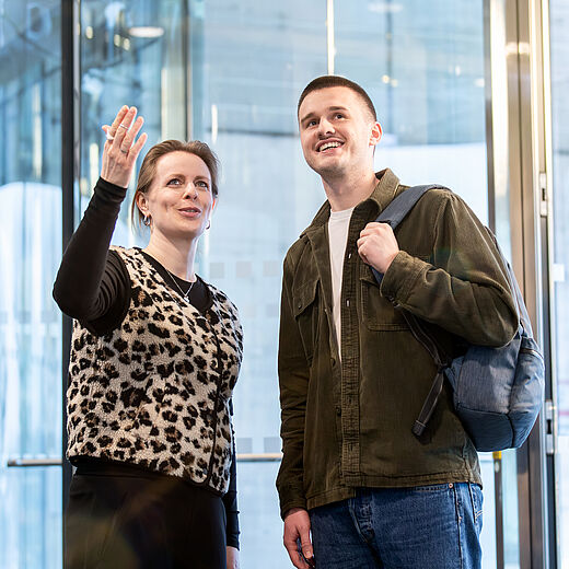 Eine Frau zeigt mit ihrer Hand in eine Richtung, während sie mit einem Mann spricht, der einen Rucksack trägt. Beide lächeln und stehen in einem modernen Gebäude mit großen Glasschiebetüren im Hintergrund.
