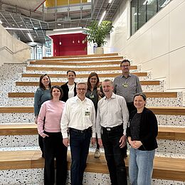 Eine Gruppe von neun Personen steht auf einer Treppe mit Holz- und Steinoptik. Sie lächeln in die Kamera. Im Hintergrund sind moderne Architektur und eine Pflanze zu sehen. Die Atmosphäre wirkt freundlich und einladend.