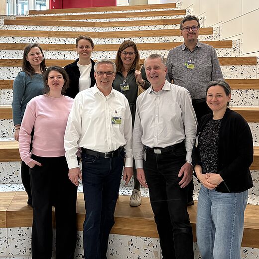 Eine Gruppe von neun Personen steht auf einer Treppe mit Holz- und Steinoptik. Sie lächeln in die Kamera. Im Hintergrund sind moderne Architektur und eine Pflanze zu sehen. Die Atmosphäre wirkt freundlich und einladend.