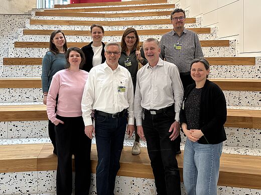 Eine Gruppe von neun Personen steht auf einer Treppe mit Holz- und Steinoptik. Sie lächeln in die Kamera. Im Hintergrund sind moderne Architektur und eine Pflanze zu sehen. Die Atmosphäre wirkt freundlich und einladend.