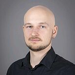 Porträt eines Mannes mit glattem Kopf und kurzem Bart. Er trägt ein schwarzes Hemd und hat einen lockeren, aber neutralen Gesichtsausdruck. Der Hintergrund ist einfarbig grau.