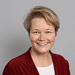 Ein lächelnde Frau mit kurzen, hellen Haaren trägt einen roten Blazer und ein weißes T-Shirt. Der Hintergrund ist einfarbig grau. Die Frau schaut direkt in die Kamera und strahlt Freundlichkeit aus.