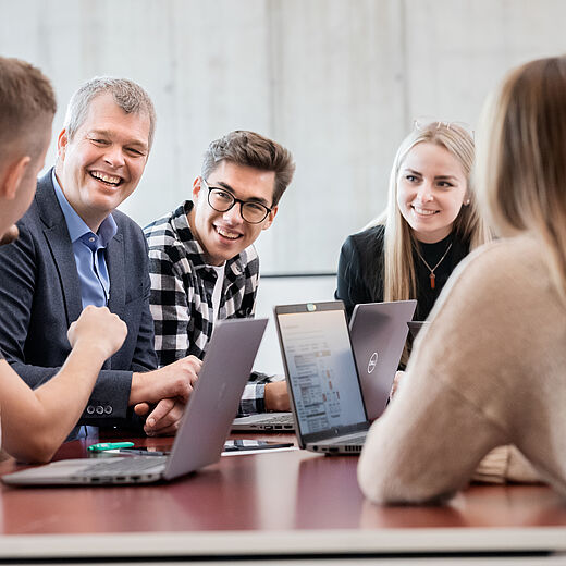 Eine Gruppe von fünf Personen sitzt an einem Konferenztisch und lächelt. Sie engagieren sich in einem Gespräch, während mehrere Laptops offen auf dem Tisch stehen. Die Atmosphäre ist freundlich und kooperativ, mit einem Fokus auf Zusammenarbeit und Austausch von Ideen.