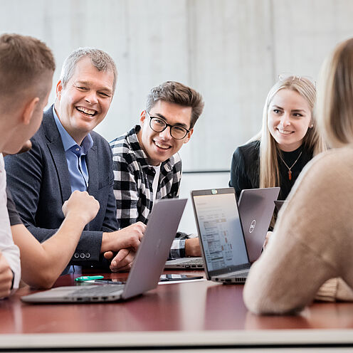 Eine Gruppe von fünf Personen sitzt an einem Konferenztisch und lächelt. Sie engagieren sich in einem Gespräch, während mehrere Laptops offen auf dem Tisch stehen. Die Atmosphäre ist freundlich und kooperativ, mit einem Fokus auf Zusammenarbeit und Austausch von Ideen.