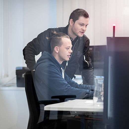 Zwei Männer sitzen an einem Schreibtisch vor Monitoren. Der Mann im Vordergrund arbeitet am Computer, während der Mann im Hintergrund über seine Schulter schaut und ihn dabei unterstützt. Die Umgebung wirkt modern und hell.