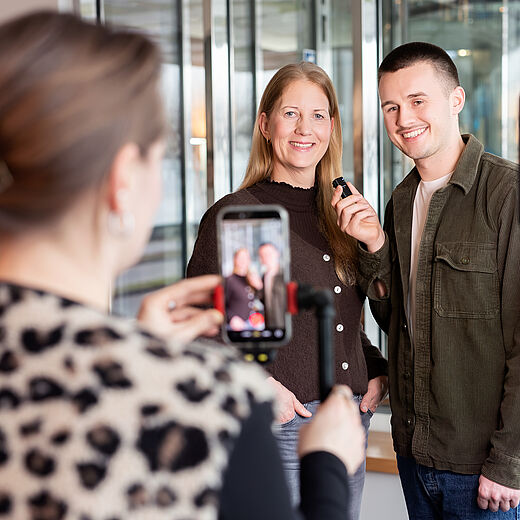 Auf dem Bild sieht man zwei Personen, eine Frau und einen Mann, die lächeln und zusammen posieren. Im Hintergrund hält eine dritte Person ein Smartphone und filmt die beiden. Sie stehen in einem hellen Raum mit großen Fenstern.