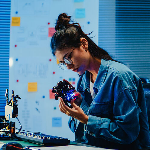 Eine Person sitzt an einem Tisch in einem modernen Arbeitsraum, betrachtet konzentriert ein elektronisches Bauteil und arbeitet daran. Um sie herum sind Werkzeuge und Geräte zur Elektronikbearbeitung sowie Computerbildschirme. Das Licht ist gedämpft, was eine kreative Atmosphäre schafft.