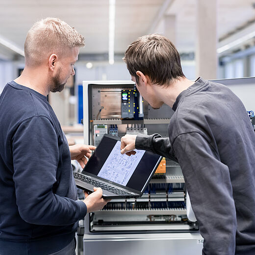 Zwei Männer stehen vor einem technischen Gerät in einer hellen Arbeitsumgebung. Einer hält einen Laptop und zeigt auf einen Plan, während der andere aufmerksam zusieht. Beide scheinen ein technisches Problem zu besprechen oder an einer Lösung zu arbeiten.