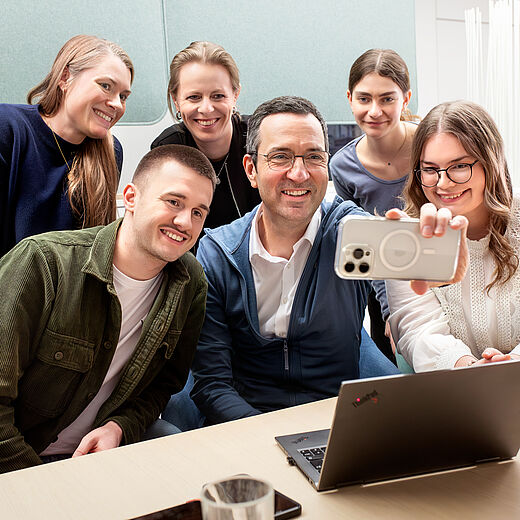 Eine Gruppe von sechs Personen steht zusammen und lächelt in die Kamera. Ein Mann in der Mitte hält ein Smartphone, um ein Selfie zu machen. Im Vordergrund steht ein Laptop auf einem Tisch. Alle scheinen fröhlich und entspannt zu sein.