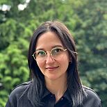 Eine junge Frau mit langen, dunklen Haaren und einer schwarzen Bluse steht lächelnd vor einem unscharfen grünen Hintergrund. Sie trägt eine modische Brille und hat einen freundlichen Ausdruck.