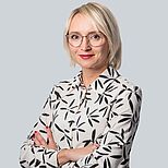 Eine Frau mit blonden, schulterlangen Haaren und Brille steht in einem neutralen Raum. Sie trägt eine Bluse mit floralem Muster und hat die Arme verschränkt. Ihr Ausdruck ist selbstbewusst und freundlich.
