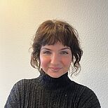 Eine junge Frau mit kurzen, gewellten braunen Haaren und einem freundlichen Lächeln steht vor einer neutralen Wand. Sie trägt einen schwarzen Pullover mit Rollkragen und blickt direkt in die Kamera.