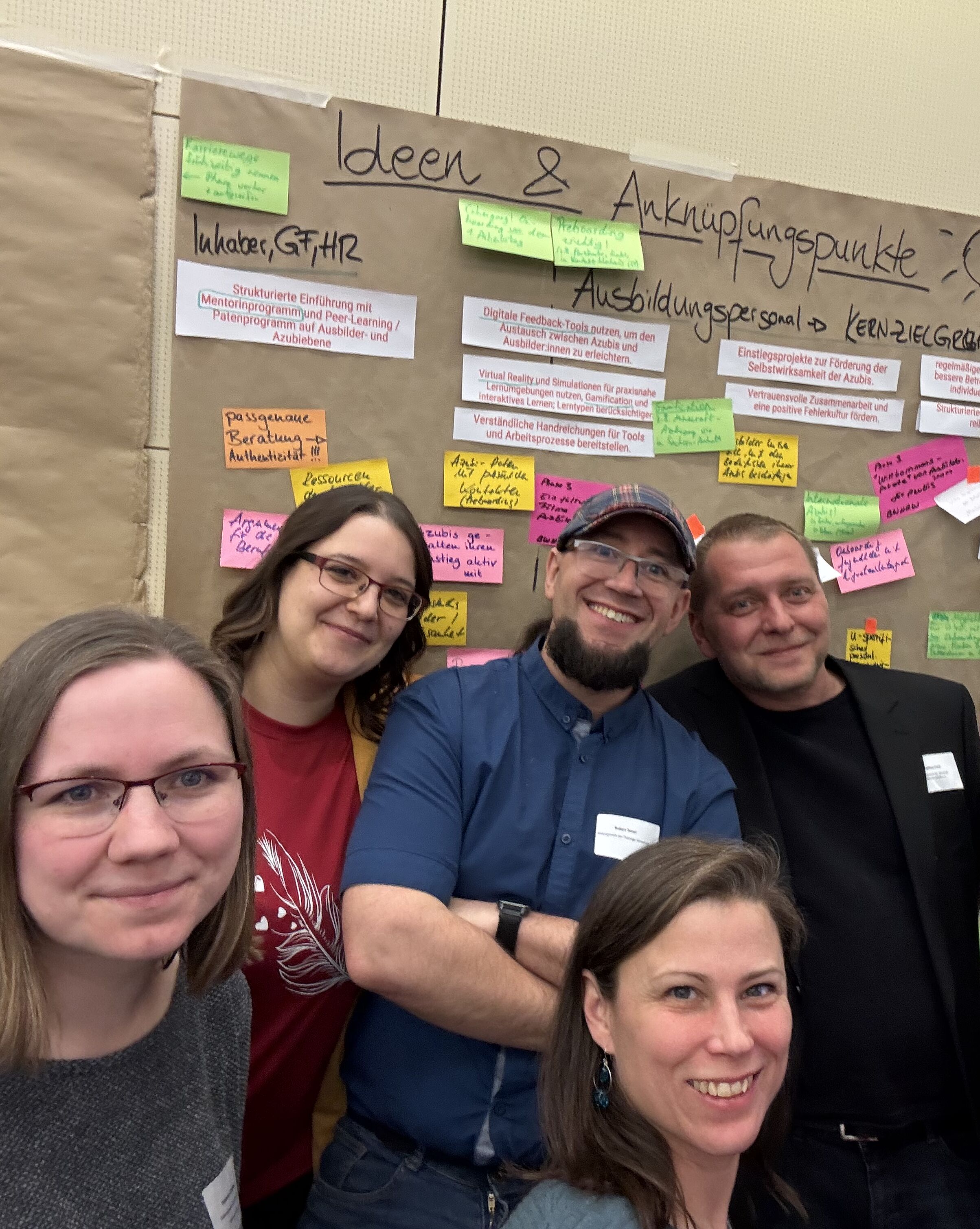Eine Gruppe von acht Personen steht lächelnd vor einem Pinnwand mit bunten Notizen. Auf der Pinnwand sind Ideen und Anknüpfungspunkte zu sehen, die sich mit Ausbildung und Zusammenarbeit befassen. Alle Teilnehmer wirken engagiert und erfreut.