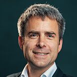 Ein lächelnder Mann mit kurzen, grauen Haaren trägt einen dunklen Anzug und ein weißes Hemd. Er steht vor einem einfarbigen Hintergrund und strahlt Freundlichkeit und Selbstbewusstsein aus.