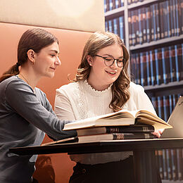 Zwei junge Frauen sitzen an einem Tisch in einer Bibliothek. Eine von ihnen sieht lächelnd auf einen Laptop, während die andere ein Buch aufgeschlagen anseht. Im Hintergrund sind Regale mit Bücher zu sehen.