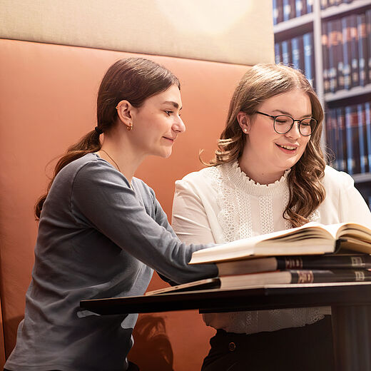 Zwei junge Frauen sitzen an einem Tisch in einer Bibliothek. Eine von ihnen sieht lächelnd auf einen Laptop, während die andere ein Buch aufgeschlagen anseht. Im Hintergrund sind Regale mit Bücher zu sehen.