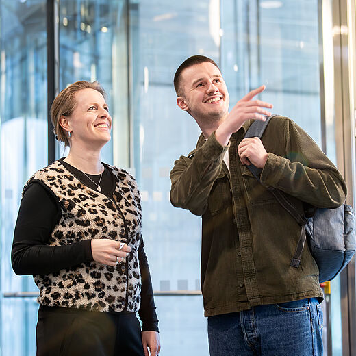 Zwei Personen stehen in einem modernen Eingangsbereich. Eine Frau mit kurzer Frisur trägt ein gemustertes Oberteil und lächelt, während ein Mann mit kurzem Haar und einem langen Oberhemd auf etwas zeigt und lächelt. Sie scheinen sich über etwas zu unterhalten.