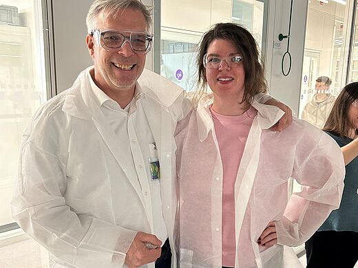 Zwei Personen stehen nebeneinander in einem Labor. Beide tragen weiße Laborkittel und Schutzbrillen. Der Mann lächelt und hält eine Hand in die Kamera. Die Frau steht lächelnd neben ihm mit einem Arm um seine Schulter. Im Hintergrund sind unscharfe Personen und Fenster sichtbar.