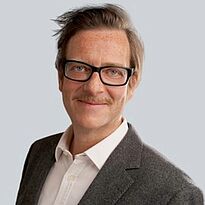 Ein lächelnder Mann mit Brille und Schnurrbart trägt einen grauen Blazer und ein weißes Hemd. Der Hintergrund ist einfarbig hellgrau. Der Mann sieht freundlich und selbstbewusst aus.