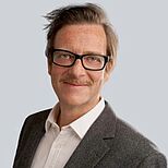 Ein lächelnder Mann mit Brille und Schnurrbart trägt einen grauen Blazer und ein weißes Hemd. Der Hintergrund ist einfarbig hellgrau. Der Mann sieht freundlich und selbstbewusst aus.