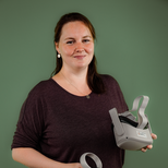 Eine Frau mit langen Haaren steht vor einem grünen Hintergrund und hält eine VR-Brille in der einen Hand und einen Controller in der anderen Hand. Sie trägt ein lockeres, dunkles Oberteil und lächelt in die Kamera.