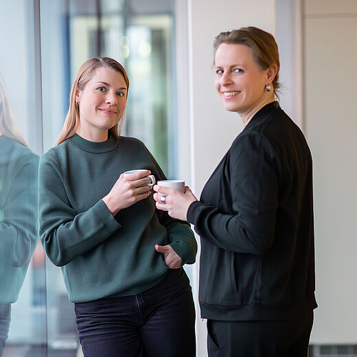 Zwei Frauen stehen an einem Fenster und lächeln. Die links stehende Frau hält einen Kaffeebecher in der Hand und trägt einen grünen Pullover. Die rechts stehende Frau hat eine dunkle Jacke an und hält ebenfalls einen Becher. Beide scheinen in einem freundlichen Gespräch vertieft zu sein.