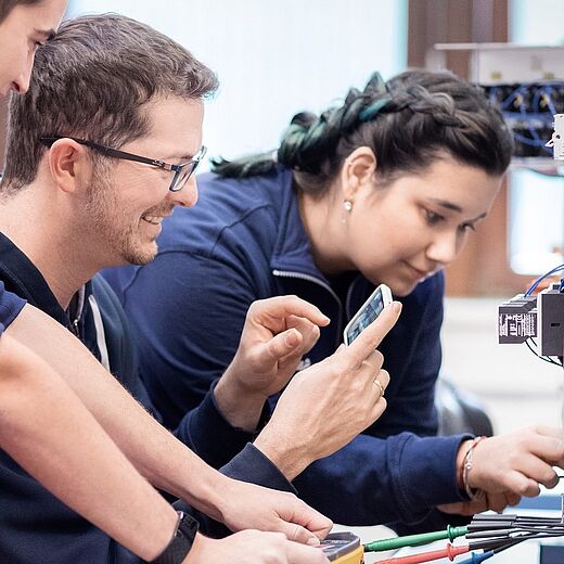 Drei Personen arbeiten zusammen an einem technischen Gerät. Einer hält ein Smartphone und sieht auf den Bildschirm, während die anderen beiden konzentriert an dem Gerät arbeiten. Sie tragen Arbeitskleidung und scheinen in einem modernen Arbeitsumfeld zu sein.