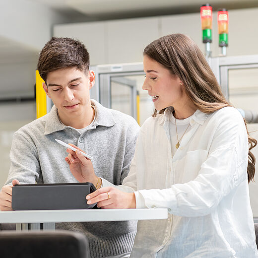Zwei Personen, ein Mann und eine Frau, sitzen an einem Tisch in einem modernen Labormilieu. Sie schauen gemeinsam auf ein Tablet, während die Frau mit einem Stift darauf zeigt. Beide wirken engagiert und in ein Gespräch vertieft.