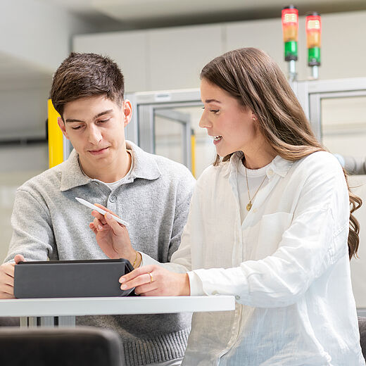 Zwei Personen, ein Mann und eine Frau, sitzen an einem Tisch in einem modernen Labormilieu. Sie schauen gemeinsam auf ein Tablet, während die Frau mit einem Stift darauf zeigt. Beide wirken engagiert und in ein Gespräch vertieft.