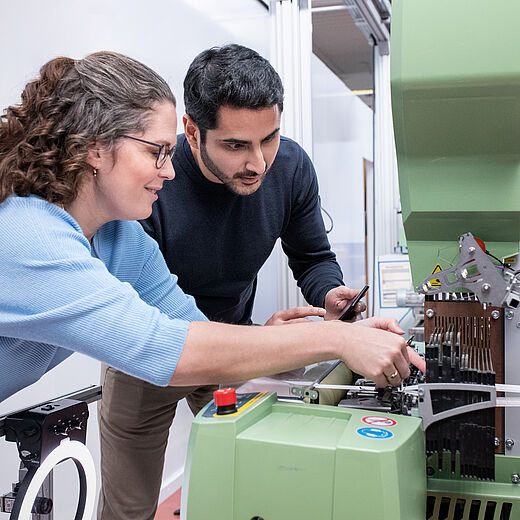 Zwei Personen, eine Frau mit lockigem Haar und Brille sowie ein Mann, arbeiten gemeinsam an einer grünen Maschine. Sie schauen konzentriert auf die Maschine und bedienen sie, wobei sie offensichtlich an einem technischen Projekt arbeiten.