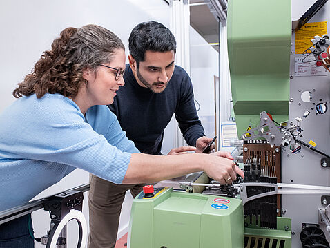 Zwei Personen, eine Frau mit lockigem Haar und Brille sowie ein Mann, arbeiten gemeinsam an einer grünen Maschine. Sie schauen konzentriert auf die Maschine und bedienen sie, wobei sie offensichtlich an einem technischen Projekt arbeiten.