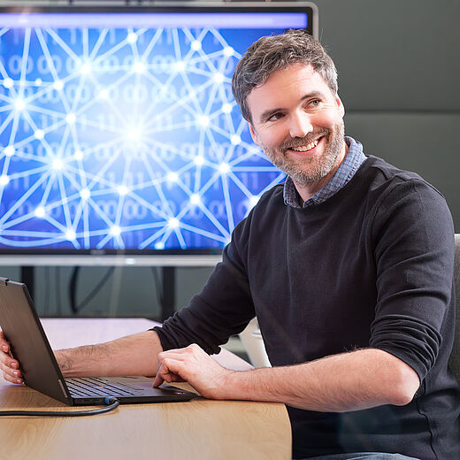 Ein lächelnder Mann sitzt an einem Tisch und arbeitet mit einem Laptop. Im Hintergrund ist ein Bildschirm mit grafischen Darstellungen von Datenverbindungen und einem Netzwerk zu sehen. Der Mann wirkt freundlich und ansprechend in seiner Umgebung.