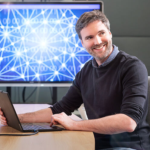Ein lächelnder Mann sitzt an einem Tisch und arbeitet mit einem Laptop. Im Hintergrund ist ein Bildschirm mit grafischen Darstellungen von Datenverbindungen und einem Netzwerk zu sehen. Der Mann wirkt freundlich und ansprechend in seiner Umgebung.