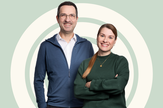Zwei Personen stehen vor einem sanften, kreisförmigen Hintergrund. Eine Frau mit langen, braunen Haaren und einem grünen Pullover lächelt freundlich. Ein Mann mit Brille und graumeliertem Haar trägt einen blauen Pullover und ebenfalls ein Lächeln. Sie strahlen Vertrauen und Offenheit aus.