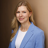 Porträt einer Frau mit blonden, langen Haaren. Sie trägt einen blauen Blazer über einem weißen T-Shirt und lächelt freundlich. Der Hintergrund ist in einem warmen Braunton gehalten.