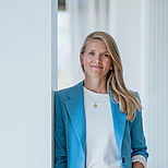 Eine lächelnde Frau mit langen, blonden Haaren steht an einer Wand. Sie trägt einen hellblauen Blazer über einem weißen T-Shirt und Jeans. Ihr Ausdruck ist freundlich und selbstbewusst, während sie entspannt mit einer Hand in der Hosentasche steht.