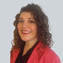 Eine lächelnde Frau mit lockigem, braunem Haar und einem roten Blazer steht vor einem hellen Hintergrund. Sie strahlt Selbstbewusstsein und Freundlichkeit aus.