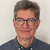 Ein Mann mit grauen, kurzen Haaren und einer Brille lächelt in die Kamera. Er trägt ein dunkles Hemd und hat einen freundlichen Gesichtsausdruck. Der Hintergrund ist hell und neutral.