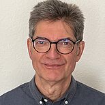 Ein Mann mit grauen, kurzen Haaren und einer Brille lächelt in die Kamera. Er trägt ein dunkles Hemd und hat einen freundlichen Gesichtsausdruck. Der Hintergrund ist hell und neutral.